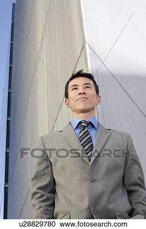 Stock Image - Young Businessman standing, hands in pockets, low angle view. Fotosearch