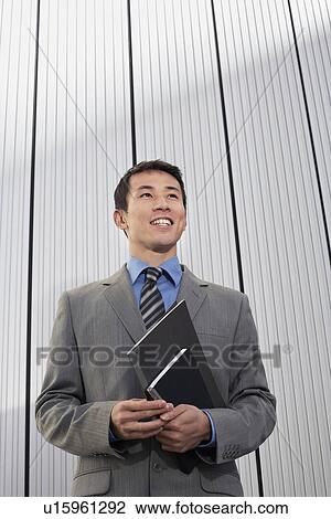 Stock Image - Young Businessman standing, holding notebook, low angle view. Fotosearch