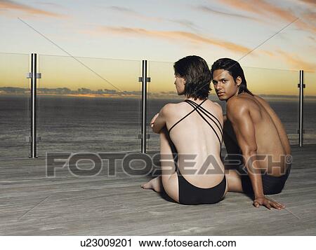 Stock Image - Young couple in bathing suits sitting on deck looking at sunset over ocean. Fotosearch