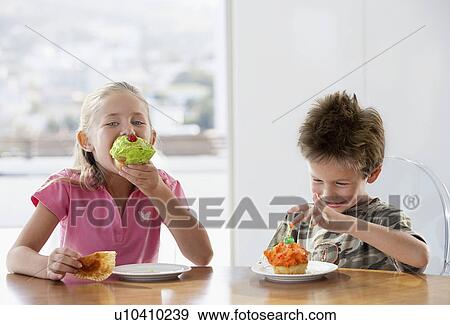 Stock Photo - Young girl and boy eating cup cakes at table, half length. Fotosearch
