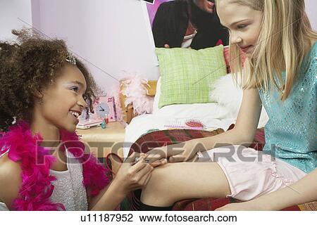 Stock Image - Young girl applying nail polish to friends finger in bedroom. Fotosearch