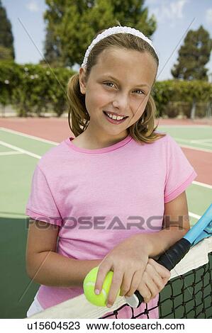 Stock Image - Young girl with tennis racket and ball by net at tennis court, portrait. Fotosearch