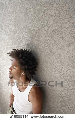 Picture - Young man leaning against wall listening to music through headphones, portrait. Fotosearch