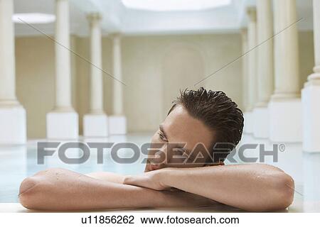 Stock Image - Young man leaning on edge of swimming pool, portrait. Fotosearch