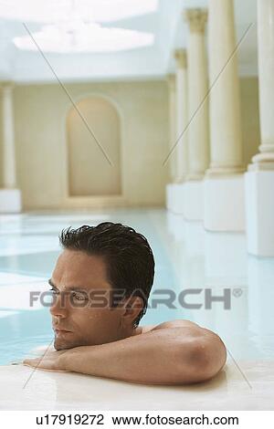 Stock Image - Young man leaning on edge of swimming pool, portrait. Fotosearch