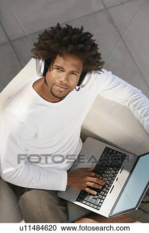Young man sitting on sofa outside using laptop and listening to music through earphones, portrait, overhead view View Large Photo Image Stock Image - Young man sitting on sofa outside using laptop and listening to music through earphones, portrait, overhead view. Fotosearch