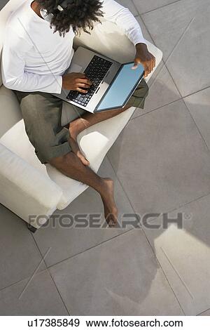 Young man sitting on sofa outside using laptop and listening to music through earphones, overhead view View Large Photo Image Stock Photo - Young man sitting on sofa outside using laptop and listening to music through earphones, overhead view. Fotosearch