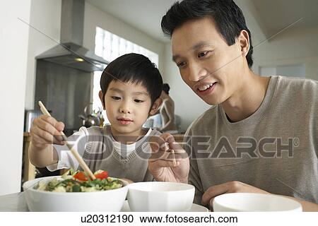 Stock Image - Young parent sitting at table, watching son trying to use chopsticks. Fotosearch