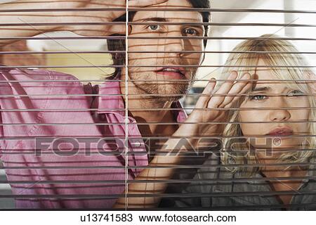 Stock Image - Young woman and man looking through Venetian Blinds, front view.. Fotosearch