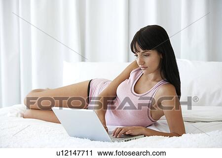Young woman lying on bed using laptop, portrait View Large Photo Image Stock Image - Young woman lying on bed using laptop, portrait. Fotosearch