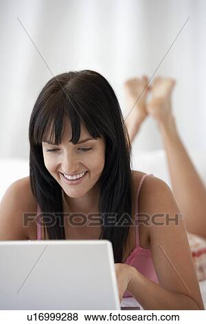 Stock Photo - Young woman lying on bed using laptop, portrait. Fotosearch