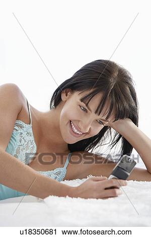 Stock Image - Young woman lying on bed using mobile phone, portrait. Fotosearch