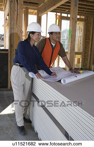 Stock Image - Architect and construction worker looking at blueprints inside half constructed house. Fotosearch