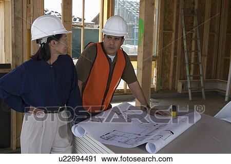 Stock Image - Architect and construction worker looking at blueprints inside half constructed house. Fotosearch