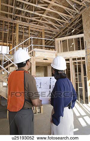 Stock Image - Architect and construction worker looking up while holding with blueprints inside half constructed house. Fotosearch