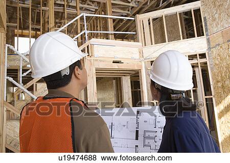 Stock Photo - Architect and construction worker looking up while holding with blueprints inside half constructed house. Fotosearch