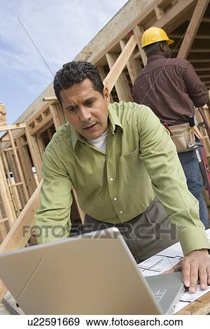Stock Photo - Architect using laptop on construction site with construction worker standing behind him. Fotosearch