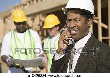 Picture - Architect using mobile phone and two construction works looking at blueprints. Fotosearch
