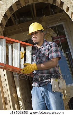 Stock Image - Construction worker carrying step ladder on construction site. Fotosearch