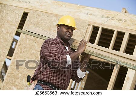 Stock Image - Construction worker carrying wooden plank. Fotosearch