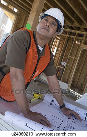 Stock Image - Construction worker examining blueprints inside half constructed house. Fotosearch