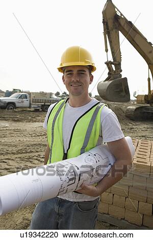 Stock Image - Construction worker holding blueprints on site, portrait. Fotosearch