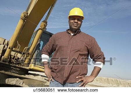 Stock Image - Construction Worker in hard hat standing, hand on hip, in front of heavy machinery. Fotosearch