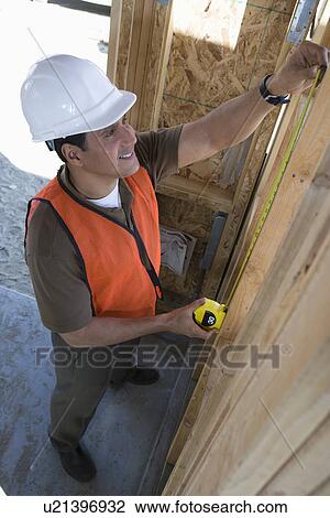 Stock Image - Construction worker measuring wall with measure taper inside half constructed house. Fotosearch
