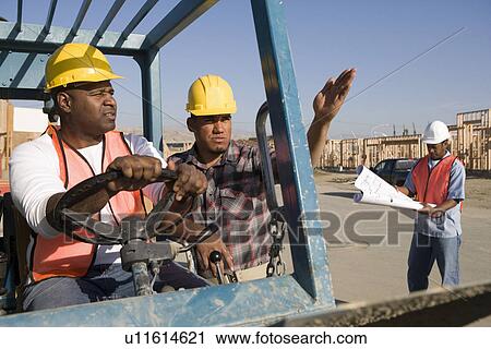 Stock Image - Construction worker showing direction to another worker steering vehicle. Fotosearch