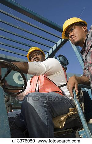 Stock Image - Construction worker sitting inside vehicle with another worker standing. Fotosearch
