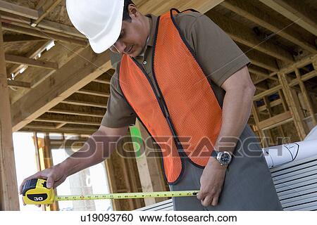 Stock Image - Construction worker using tape measure inside half constructed house. Fotosearch