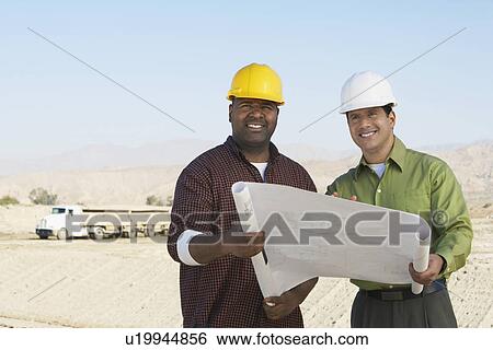 Stock Photograph - Construction Workers wearing hard hats, looking at plan on Site. Fotosearch