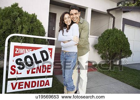 Stock Image - Couple standing in front of house with Sold sign, portrait. Fotosearch