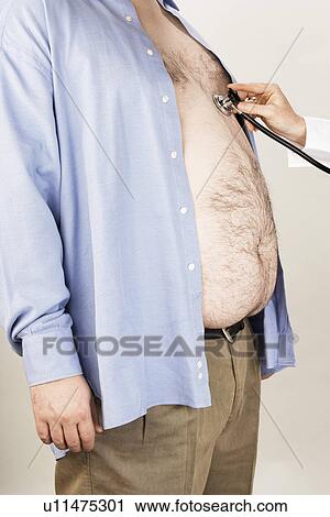 Stock Image - Doctor hand with stethoscope on overweight man's heart, side view, mid section. Fotosearch