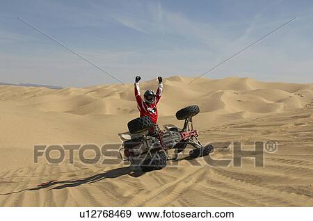 Stock Photo - Driver cheering by tipped-over quad bike in desert. Fotosearch