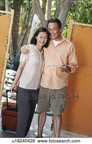 Stock Photo - Holidaymaker couple standing at gate holding suitcase and brochure, (portrait). Fotosearch