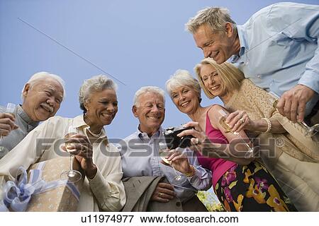 Stock Photo - Low angle view of senior woman showing pictures on digital camera to her friends. Fotosearch