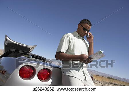 Stock Image - Man using cell phone and map by sports car at side of road. Fotosearch