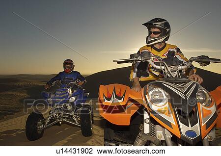 Stock Image - Men sitting on quad bikes in desert at sunset, close up. Fotosearch