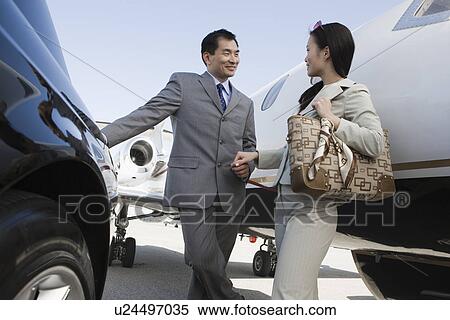 Stock Photography - Mid-adult Asian businesswoman and businessman flirting outside of car and airplane.. Fotosearch