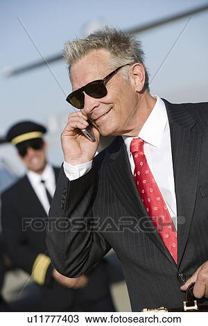 Stock Image - Mid-adult businessman talking on phone, airline pilot standing in background.. Fotosearch