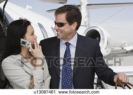 Stock Image - Mid-adult businesswoman and businessman flirting in front of airplane.. Fotosearch