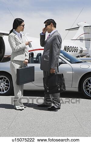 Stock Image - Mid-adult businesswoman and mid-adult businessman standing in front of convertible and talking.. Fotosearch