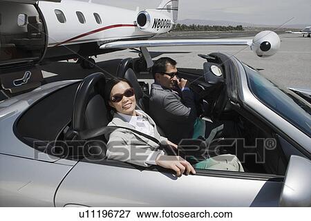 Stock Photo - Mid-adult businesswoman and mid-adult businessman sitting in convertible on landing strip.. Fotosearch