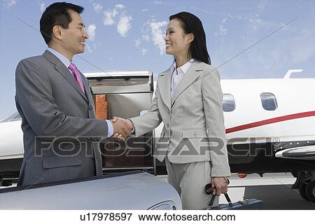 Stock Photo - Mid-adult businesswoman and mid-adult businessman shaking hands in front of private plane.. Fotosearch