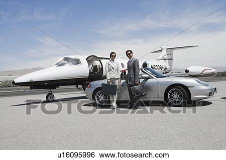 Stock Photograph - Mid-adult businesswoman and mid-adult businessman standing in front of convertible and private jet on landing strip.. Fotosearch