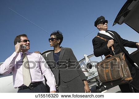 Stock Image - Mid-adult businesswoman, businessman and chauffeur in front of airplane on runway, low angle view.. Fotosearch