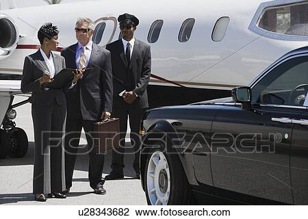 Stock Image - Mid-adult businesswoman, senior businessman and chauffeur in front of private airplane.. Fotosearch