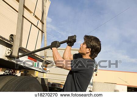 Stock Image - Mid-adult man adjusting strapping of truck loaded with wood. Fotosearch