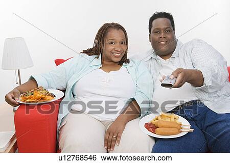 Stock Photograph - Mid-adult overweight couple sitting on sofa with meal and watching television. Fotosearch